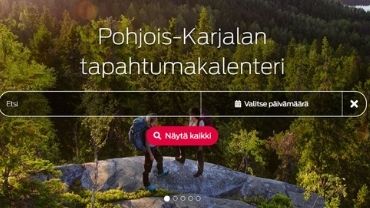 Näkymä tapahtumakalenterin etusivulta, jossa kaksi naista seisoo selin kuvaajaan ja katsoo Kolin kallioilta aukeavaa metsäistä järvimaisemaa.