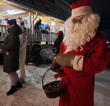 Kuvassa joulupukki ulkotiloissa. Kuva otettu viime vuoden joulukuussa Sovintolan edustalla Kesälahdella. Joulupukilla on kori kädessään ja taustalla näkyy ihmisiä, samoin Sovintolan rakennus.